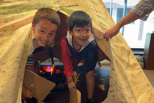 Two boys holding pencils and clipboards look out from inside a tipi at the History Museum.